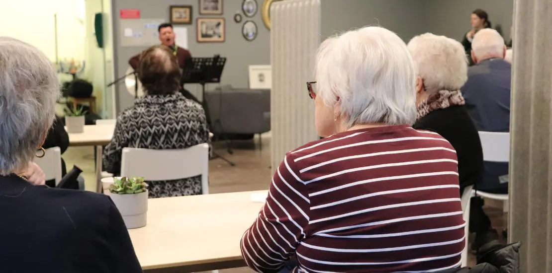 Äldre personer sitter i publik, sångare med gitarr på scen.