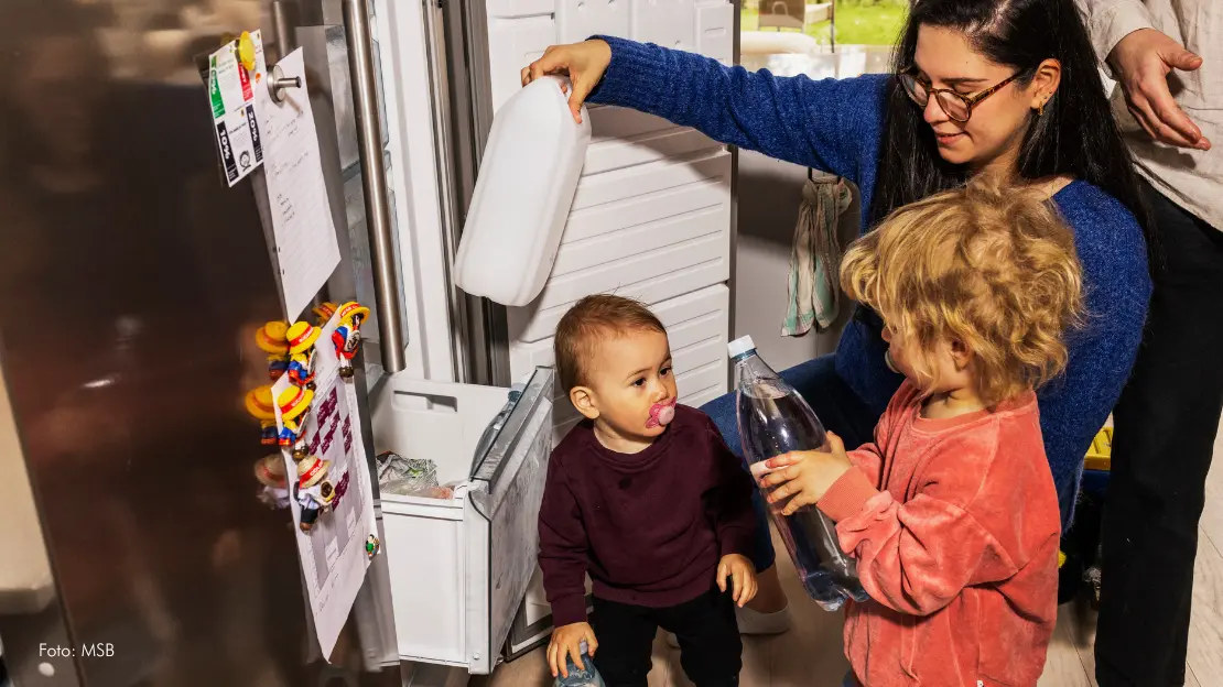 Kvinna och två små barn står framför frysen och håller mjölk och dricksvatten.