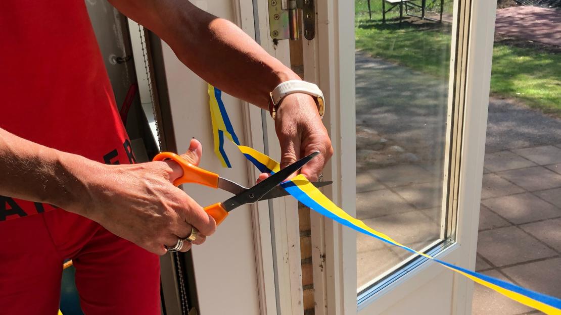 Händer håller i en orange sax och klipper ett blå-gult band i en dörröppning.