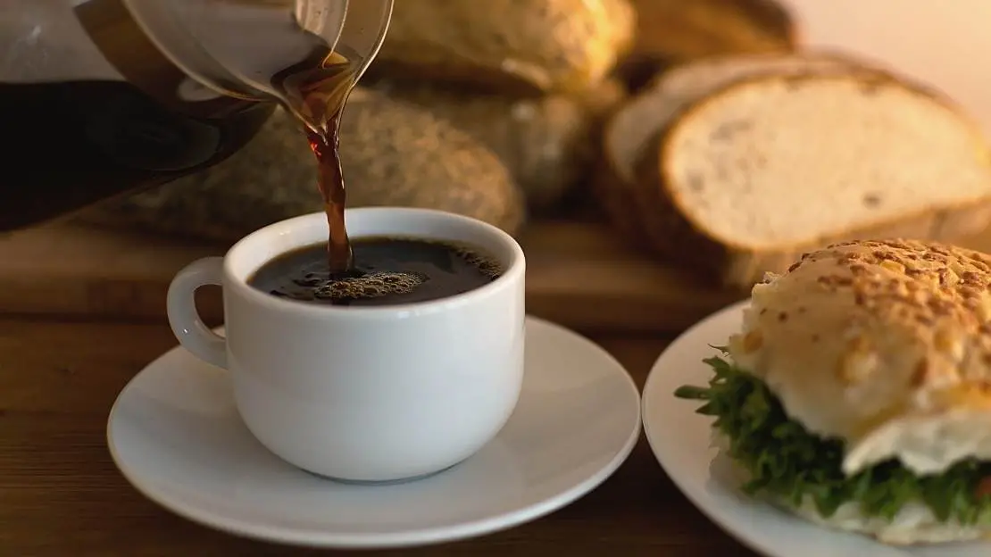 Kopp kaffe hälls upp, bredvid och i bakgrunden finns frallor. 