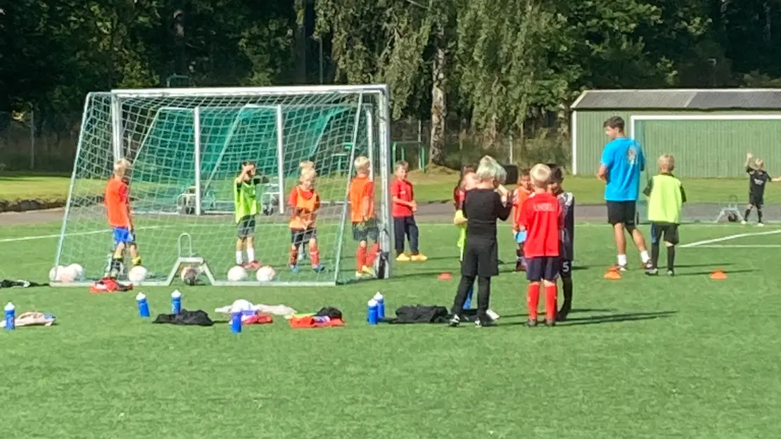 Flera barn står vid ett fotbollsmål och har träning med en ungdom som ledare.