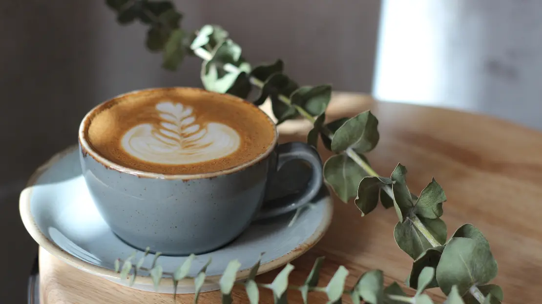 ljusblå kaffekopp på matchande fat står på litet bord med en eukalyptus kvist