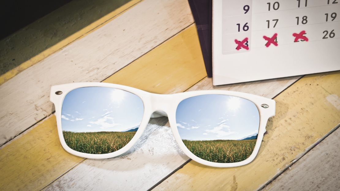 Solglasögon speglar ett soligt landskap. I bakgrunden syns kalender med några genomstrukna datum.