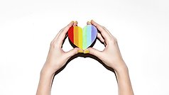 en person håller ett litet hjärta i regnbågsfärgerna upp mot en vit vägg. 
