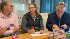 Tre personer sitter runt ett bord och pysslar. En person tar sig om kroppen då den fryser.