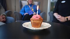 I försgrunden syns en cupcake med rosa frosting och ett ljus samt EU-flagga och Sverige flagga i, tre personer sitter i bakgrunden.