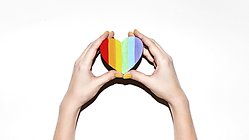 en person håller ett litet hjärta i regnbågsfärgerna upp mot en vit vägg. 